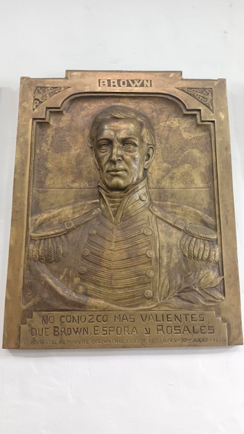 Placa de bronce rectangular con relieve de un busto de hombre en uniforme militar, texto superior "BROWN" y cita sobre el Combate de Quilmes