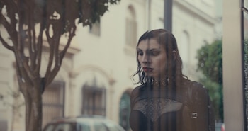 Una persona con ropa negra, gargantilla metálica y maquillaje oscuro reflejada en un cristal en la calle