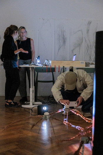 Tres personas en una galería oscura. Dos mujeres hablan junto a una mesa con un microscopio y una caja. Un hombre agachado toma fotos de una escultura con luz