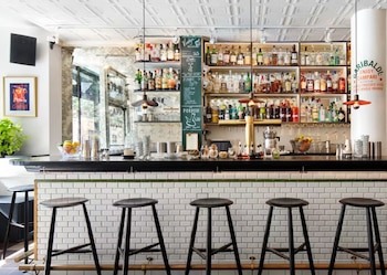 Vista frontal de un bar con una barra negra, taburetes negros, una pared de azulejos blancos y estantes llenos de botellas de licor y equipo de coctelería