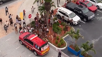 Vista aérea de un grupo de personas en una calle con un vehículo de emergencia rojo, varios coches estacionados, palmeras y una señal de alto.