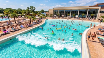 Piscina de olas al aire libre con agua turquesa, personas nadando y flotando, bañistas tomando el sol en tumbonas, palmeras y un edificio moderno al fondo.