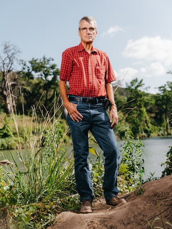 Hombre de cabello gris y gafas con camisa de cuadros roja, jeans, cinturón negro y zapatos marrones, de pie junto a un río con vegetación