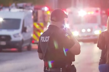 Oficial de policía de espaldas con uniforme que dice "POLICE". Al fondo, vehículos de emergencia, ambulancia, camión de bomberos y furgoneta, con luces encendidas