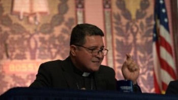 Papa Leão XIV, usando sandálias e óculos, gesticulando com as mãos levantadas, sentado em frente a uma parede estampada e uma bandeira americana