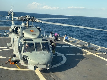 Un helicóptero militar posado en la cubierta de un barco con tripulantes en la pista y la costa en el horizonte