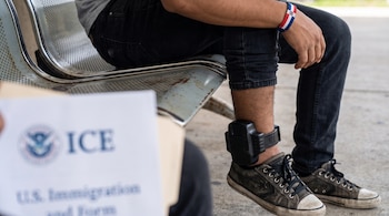 Pierna de un joven con grillete electrónico negro, zapatillas oscuras y pulsera dominicana. Documento borroso con el logo de ICE en primer plano.