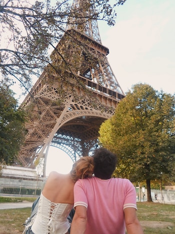 Una mujer de espaldas apoyada sobre el hombro de un hombre mientras observan la Torre Eiffel desde un parque