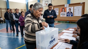 Mujer de cabello gris y suéter a rayas deposita un voto en una urna transparente de la ONPE en un centro de votación. Personas en fila y miembros de mesa también son visibles.