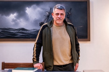 Hombre de pie con chaqueta deportiva y camiseta beige frente a un cuadro con un barco en un mar de oleaje oscuro.