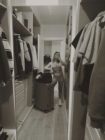 Mujer rubia en vestidor frente a un espejo, con ropa deportiva, toma selfie con móvil, junto a maleta gris y bolso negro
