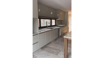 Cocina moderna con encimera de mármol, fregadero bajo una ventana, muebles de color gris claro, piso de piedra y apliques de luz en la pared