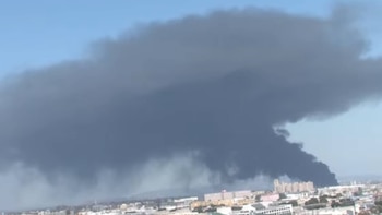 Una enorme columna de humo oscuro se eleva desde el horizonte de una ciudad con edificios visibles bajo un cielo mayormente azul