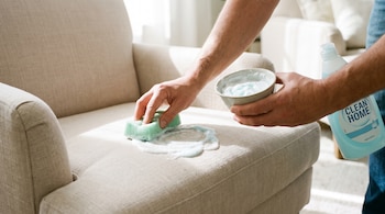 Manos aplican una mezcla espumosa con una esponja verde sobre la tapicería de un sillón claro. Hay una botella de detergente azul.