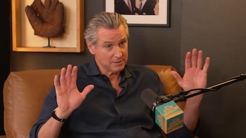 Gavin Newsom sentado en un sillón de cuero frente a un micrófono con las manos abiertas
