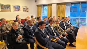 Un grupo de aproximadamente veinte personas, la mayoría hombres y una mujer, sentadas en filas en un salón, atentas a una presentación. Visten ropa de negocios