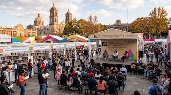 Personas caminando entre estantes llenos de libros y público sentado frente a un escenario al aire libre con una catedral de fondo.