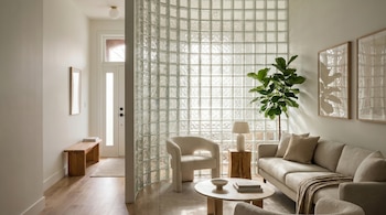 Vista de una sala de estar moderna con una pared curva de bloques de vidrio, un sofá beige, una butaca blanca, una mesa de centro y una planta grande.