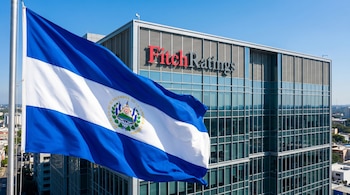 Vista aérea de la bandera de El Salvador ondeando en primer plano, con el edificio de Fitch Ratings en el fondo bajo un cielo azul claro y soleado.