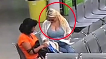 Una mujer con cabello rubio, top azul y cárdigan blanco, sentada y con la vista hacia su regazo, acompañada por otra mujer de cabello oscuro. Un círculo rojo la resalta