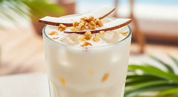 Vaso de agua fresca de coco y nuez, con hielo, trozos de coco blanco y nueces picadas flotando en la superficie, sobre un fondo borroso de playa.