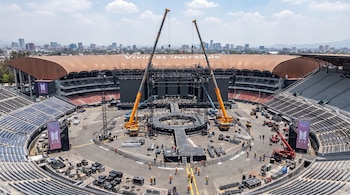 Vista aérea de un estadio con asientos vacíos, donde grúas y trabajadores construyen un gran escenario circular 360 grados con estructuras metálicas.