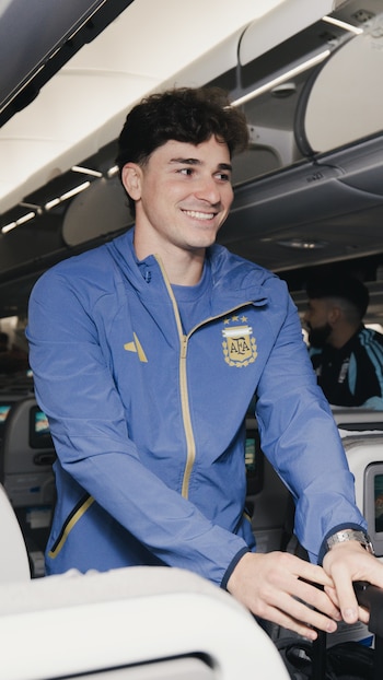 Un jugador de la selección argentina, vestido con campera deportiva azul con el escudo de AFA, sonríe dentro de un avión