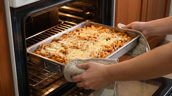 Manos sujetan con agarraderas una bandeja metálica con pasta corta cubierta de queso derretido y la colocan dentro de un horno abierto.