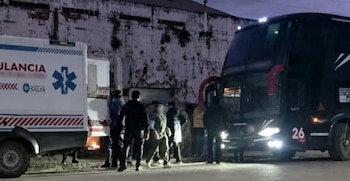 Ambulancia blanca, autobús oscuro con el número veintiséis, y varias personas junto a un edificio deteriorado en la oscuridad
