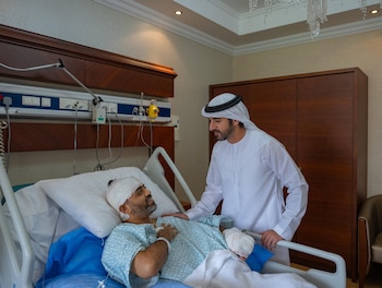 Un hombre con vestimenta emiratí conversa con un paciente vendado en la cabeza y el brazo que permanece recostado en una cama de hospital