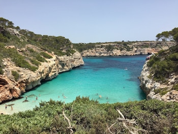Cala con aguas turquesas y azules, rodeada de acantilados rocosos con vegetación. Varias personas nadan en la orilla. Cielo despejado
