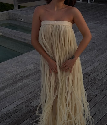 Sofía Gonet con un vestido largo color crema sin tirantes, hecho con múltiples tiras verticales, de pie en una terraza de madera junto a una piscina