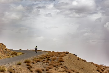 Un ciclista visto desde atrás circula por una curva de una carretera asfaltada en una colina desértica
