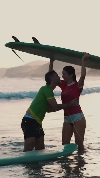 Hombre y mujer abrazados en el mar con tabla de surf sobre sus cabezas. Hombre con camiseta verde, mujer con camiseta rosa. Segunda tabla de surf en el agua