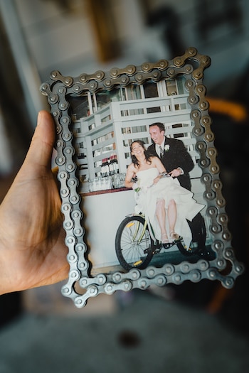 Mano sostiene un marco de fotos de cadena de bicicleta. Dentro, foto de pareja de boda sonriente en bicicleta. Botellas de vino al fondo