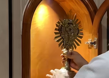 Un obispo vestido de blanco eleva una custodia de metal dorado frente a un grupo de personas