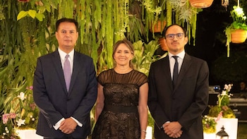 Tres personas, dos hombres y una mujer, de pie y vestidos formalmente, sonríen frente a una pared de follaje verde con plantas colgantes