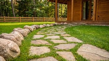 Un sendero de losas de piedra irregular sobre césped verde, bordeado por rocas, conduce a una cabaña de madera. Al fondo hay un bosque y una valla de madera.