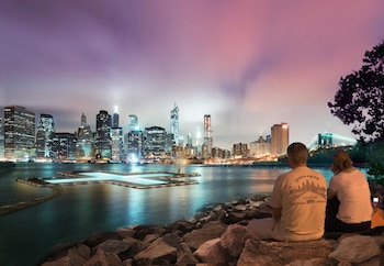 Paisaje nocturno de Nueva York: piscina flotante iluminada en el East River, rascacielos al fondo y dos personas sentadas en rocas contemplando la vista