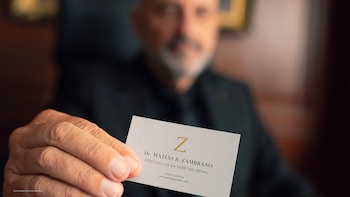 Una mano de hombre sosteniendo una tarjeta de visita blanca con letras negras que dice "Dr. Matías R. Zambrano, Especialista en Derecho Penal"; una persona difuminada se ve al fondo