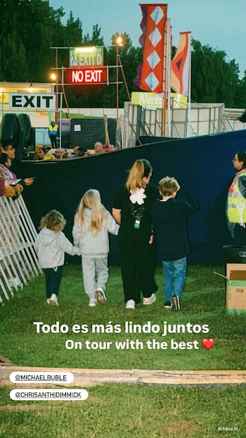 Mujer de espaldas con tres niños tomados de la mano, caminando sobre pasto frente a una barrera de tela y un escenario de festival con luces y banderas