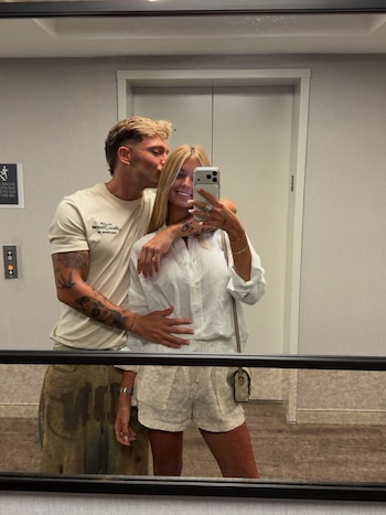 Un hombre rubio con barba besa la mejilla de una mujer rubia que toma una selfi frente al espejo de un ascensor. La mujer sonríe y sostiene un teléfono móvil