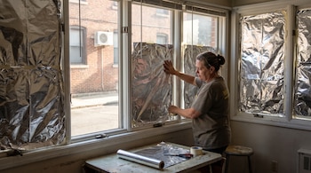 Una mujer de cabello recogido coloca papel de aluminio en varias ventanas. Un rollo de papel, tijeras y cinta adhesiva están sobre una mesa.