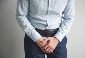 Un hombre de camisa azul y pantalón oscuro se para con las manos cruzadas sobre la ingle, frente a una pared clara