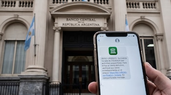 Una mano sostiene un teléfono móvil con pantalla rota mostrando un mensaje de texto fraudulento; de fondo, el edificio del Banco Central de la República Argentina.