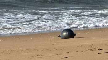 Objeto esférico de color oscuro semienterrado en la arena, con olas rompiendo en la orilla de una playa