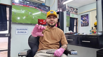 Hombre con gorra amarilla y guantes rosados sostiene tijeras y peine en un sillón de barbería frente a un televisor con fútbol.