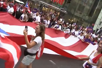 Una multitud de personas desfila en una calle de la ciudad, llevando una enorme bandera de Puerto Rico. Una mujer en primer plano sonríe mientras toma una foto