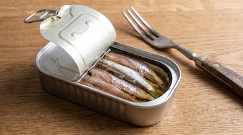 Una lata de conservas metálica abierta contiene anchoas en aceite, y a su lado hay un tenedor de metal con mango de madera, sobre una superficie de madera.