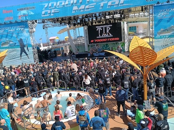 Público numeroso asiste a un concierto de heavy metal en la cubierta de un crucero, con un escenario y pantalla grande, y un jacuzzi con bañistas